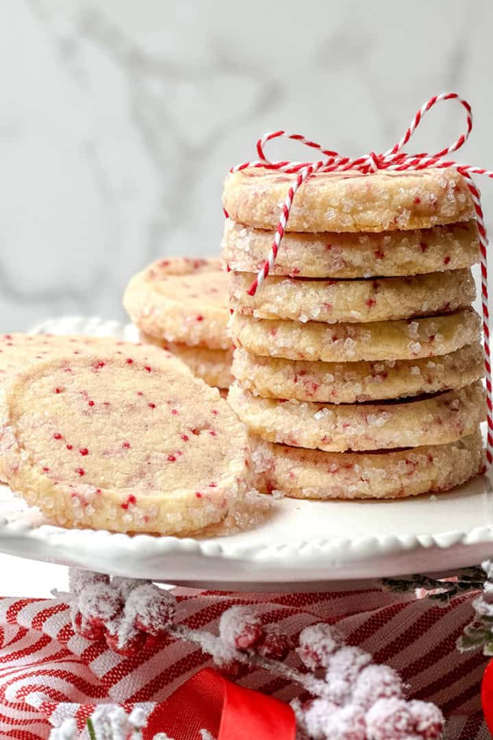 Square image of stacked Christmas slice and bake cookies with red sprinkles on a dessert stand tied with a red and white ribbon.