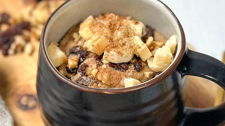 Side three-quarter view of ready to eat 2 Minute Banana Bread Mug Cake with chopped bananas, raisins, walnuts, and a sprinkling of brown sugar.