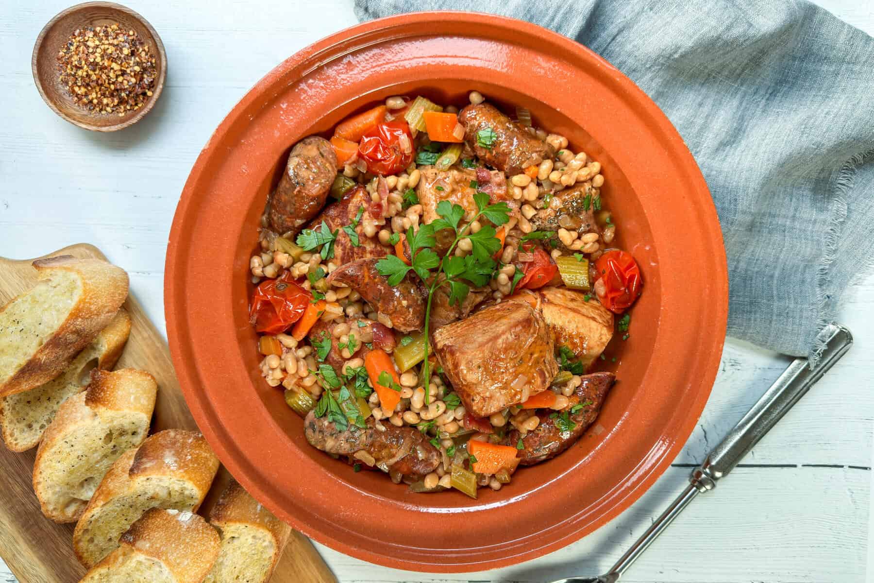 Top view of Cassoulet with fresh parsley sprigs in a terra cotta serving bowl with fresh bread slices on the side.