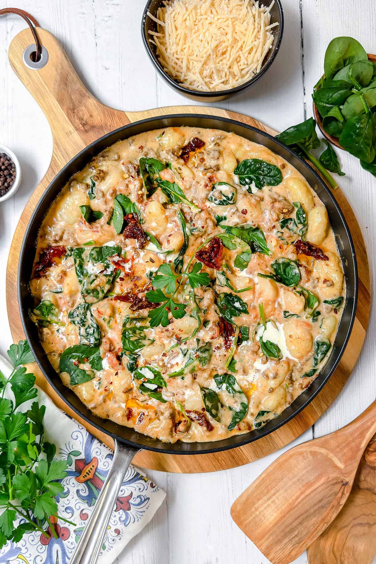 A skillet filled with creamy gnocchi recipe featuring chicken, spinach, and sun-dried tomatoes, garnished with parsley, sits on a wooden board beside a bowl of grated cheese and fresh spinach leaves.