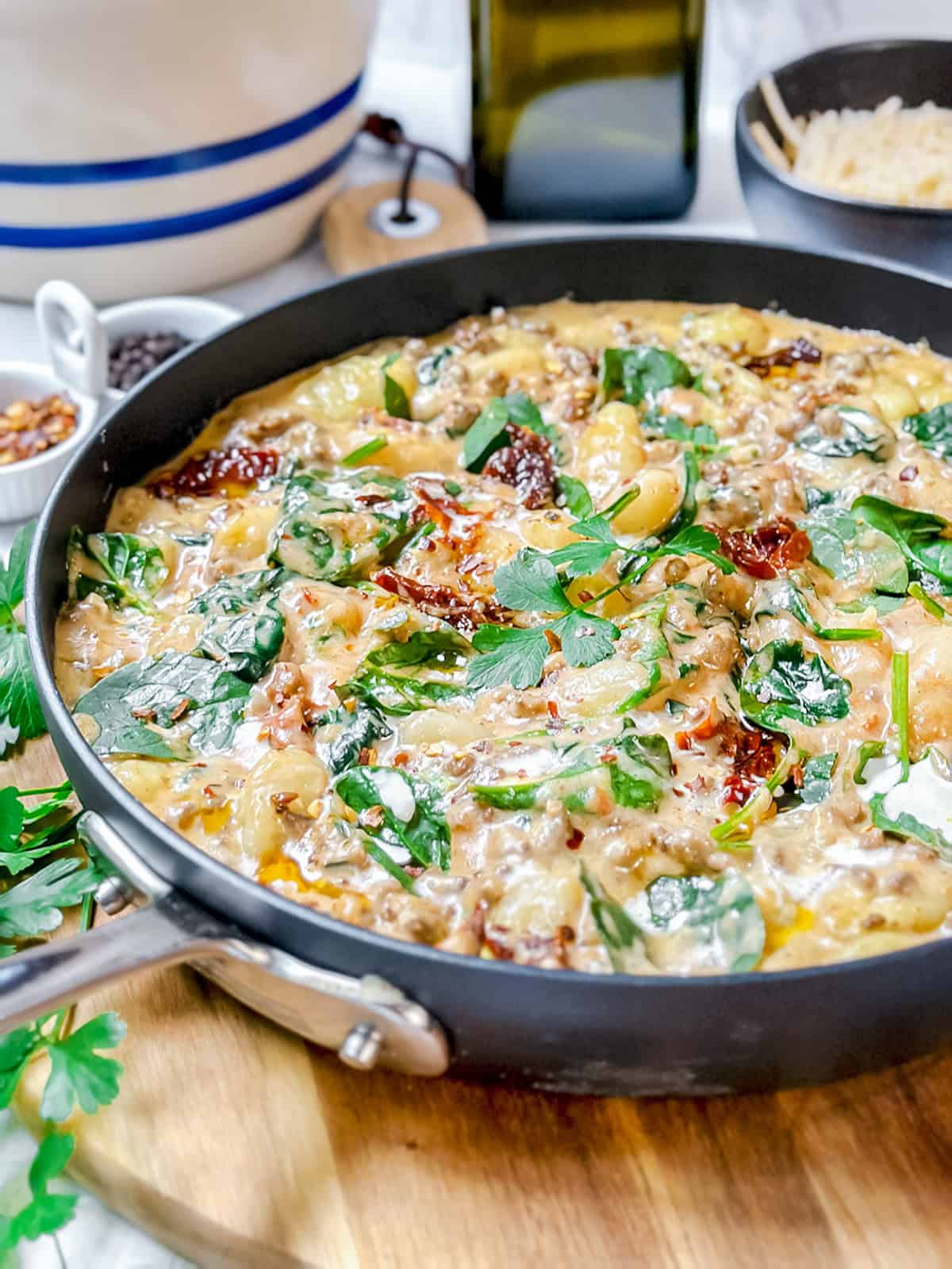 A skillet filled with creamy pasta mixed with spinach, sun-dried tomatoes, herbs, and sauce&mdash;perfect for trying your favorite gnocchi recipe&mdash;placed on a wooden board with ingredients visible in the background.