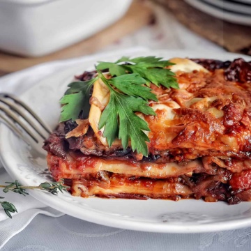 A slice of eggplant lasagna with layers of tender pasta, savory sauce, and melted cheese garnished with fresh parsley, served on a white plate with a fork beside it.