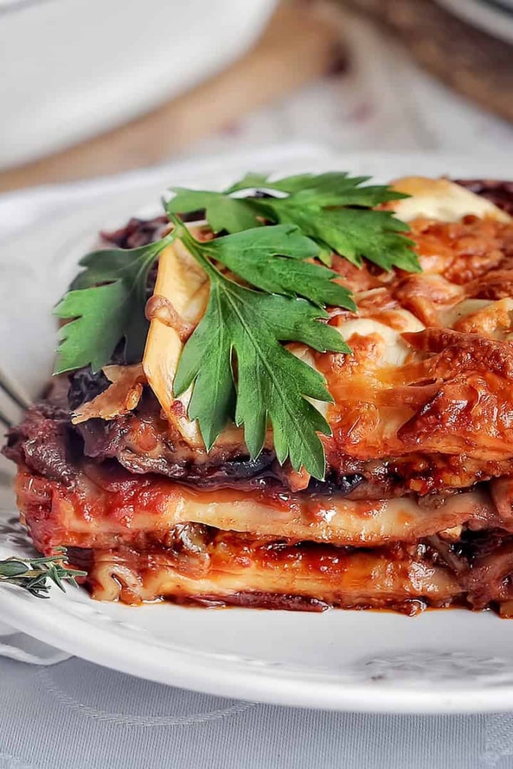 A slice of eggplant lasagna with layers of tender pasta, savory sauce, and melted cheese garnished with fresh parsley, served on a white plate with a fork beside it.