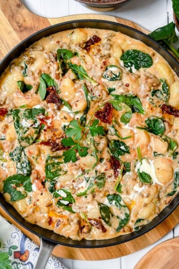 Creamy gnocchi with spinach, sun-dried tomatoes, and herbs served in a skillet on a wooden board-this irresistible gnocchi recipe is surrounded by fresh greens and seasoning bowls.