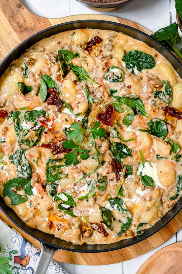 Creamy gnocchi with spinach, sun-dried tomatoes, and herbs served in a skillet on a wooden board—this irresistible gnocchi recipe is surrounded by fresh greens and seasoning bowls.