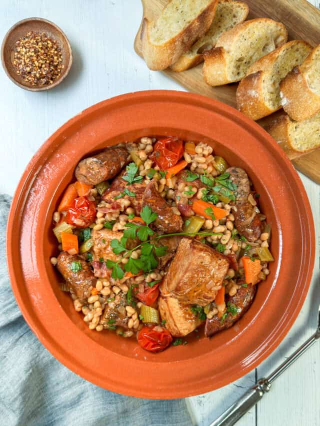 Top vertical view of slow cooker cassoulet in an earthenware pot with fresh herbs, sausages, pork, vegetables and carrots.