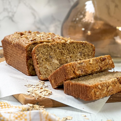 Side view of sliced Oat Flour Banana Bread on a wood cutting board with a yellow and white kitchen towel.