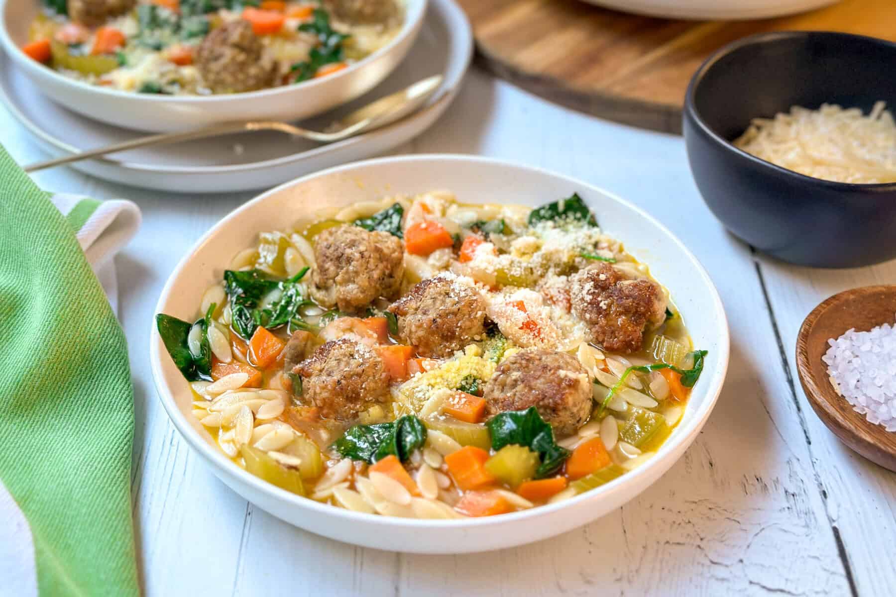 Three quarter view of Italian Wedding Soup in a white bowl with fresh Parmesan cheese and baby greens.