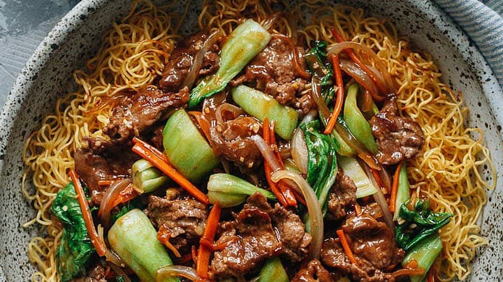 A bowl of crispy Chinese New Year noodles topped with stir-fried beef, bok choy, carrots, and onions, served with sauces and utensils on the side.