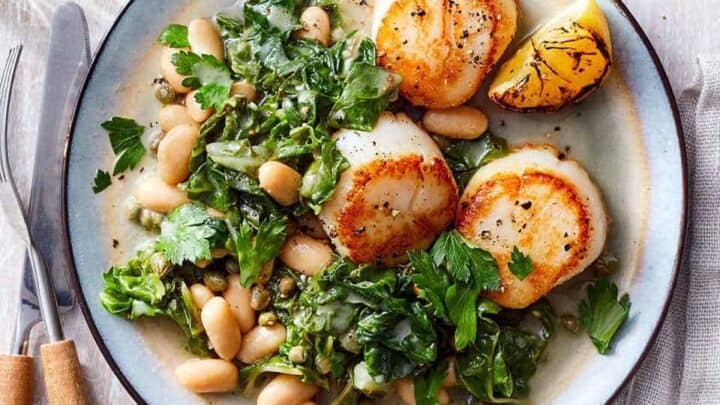 Top view of seared scallops with greens and white beans on a blue plate with wooden utensils on the side.