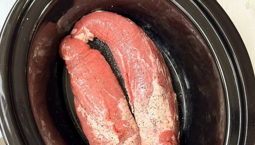 Top view of uncooked pork tenderloin, seasoned with salt and pepper, in a black crockpot insert.