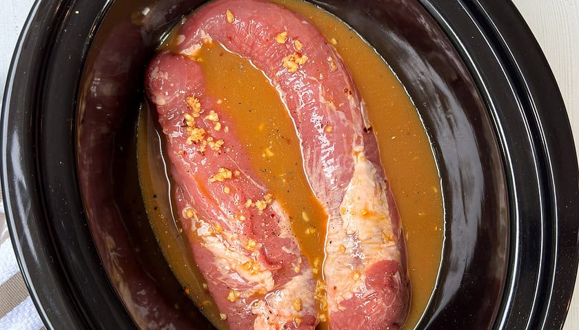 Top view of uncooked pork tenderloin with orange ginger sauce poured over in a black slow cooker insert.