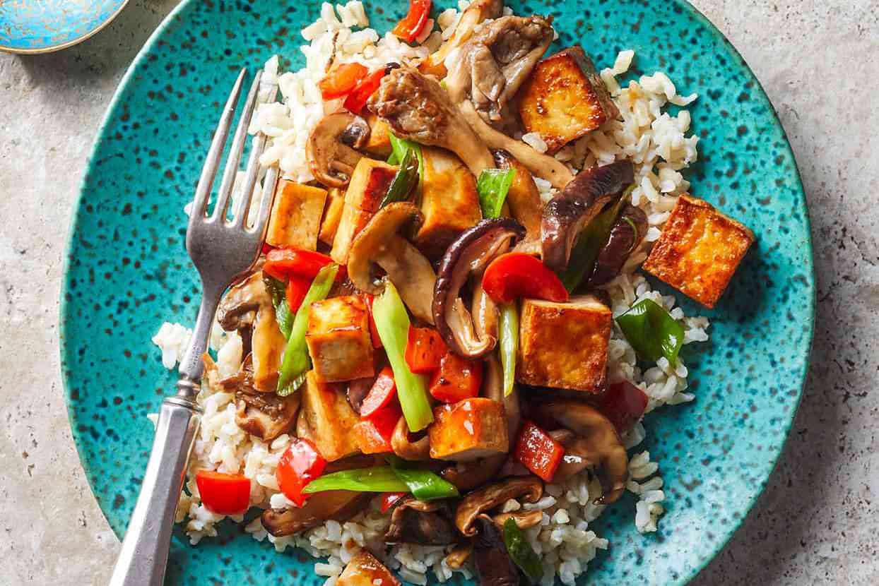 Top closeup view of tofu and mushroom stir fry over white rice with a silver fork in a blue bowl.