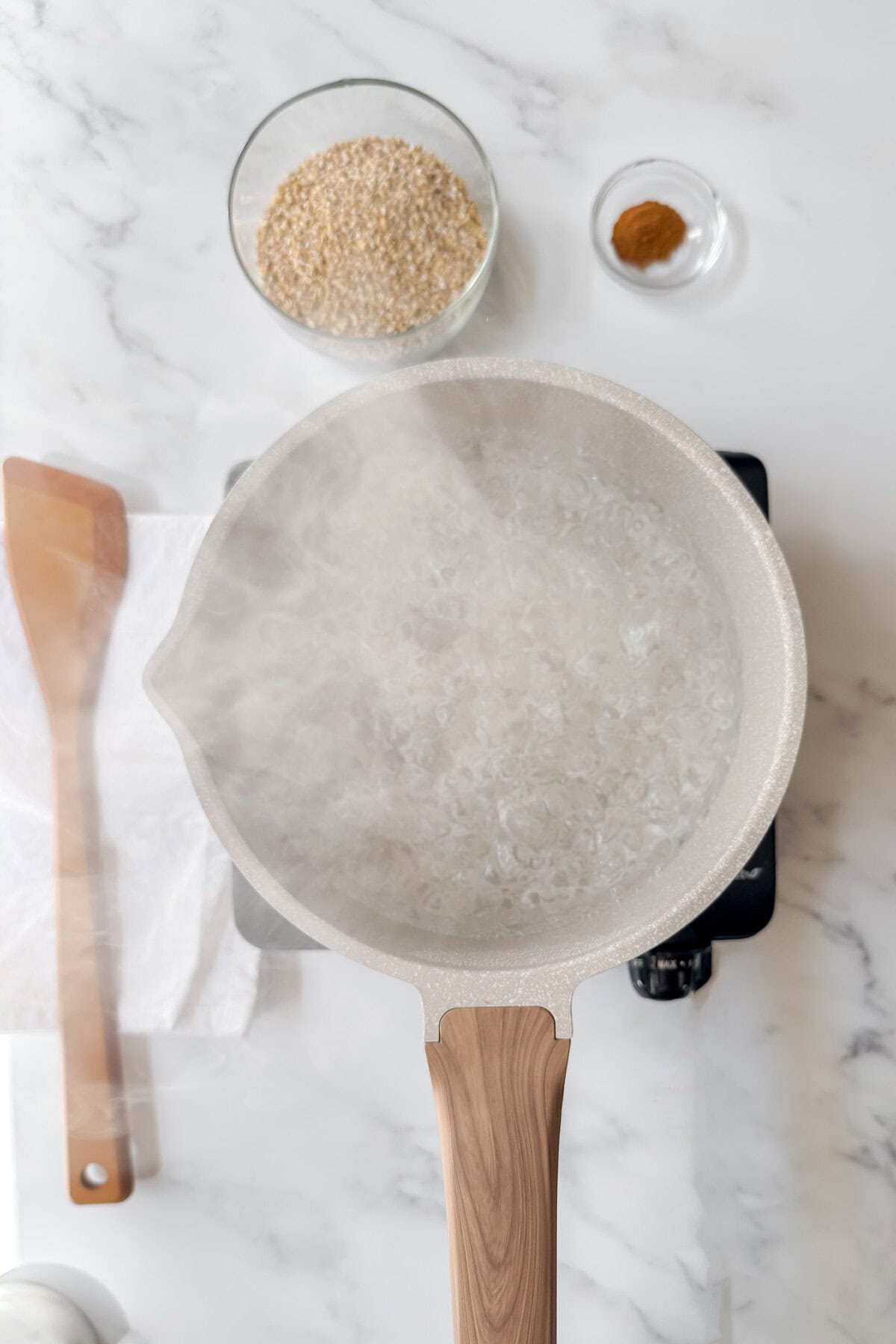 A pot of boiling water sits on a stove, releasing steam. Nearby on the marble counter are a wooden spatula, a white cloth, a bowl of Irish porridge oats, and a small dish of cinnamon.