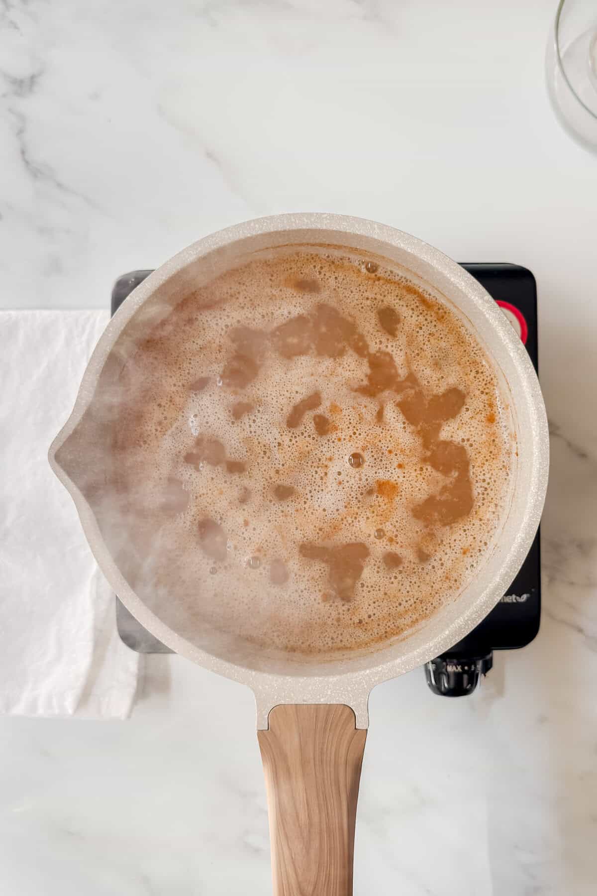 A pot with a light wooden handle is boiling Irish porridge on a black electric burner, steam rising and bubbles forming. A white cloth rests beside the pot on a marble countertop.