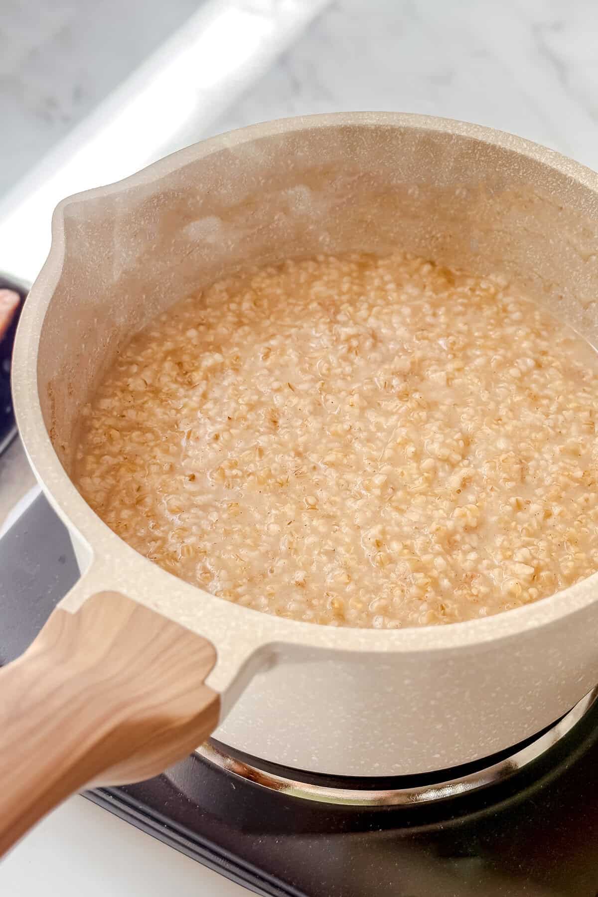 A beige saucepan with a wooden handle sits on a stovetop, filled with simmering Irish porridge. The oatmeal mixture appears thick and creamy as it cooks.