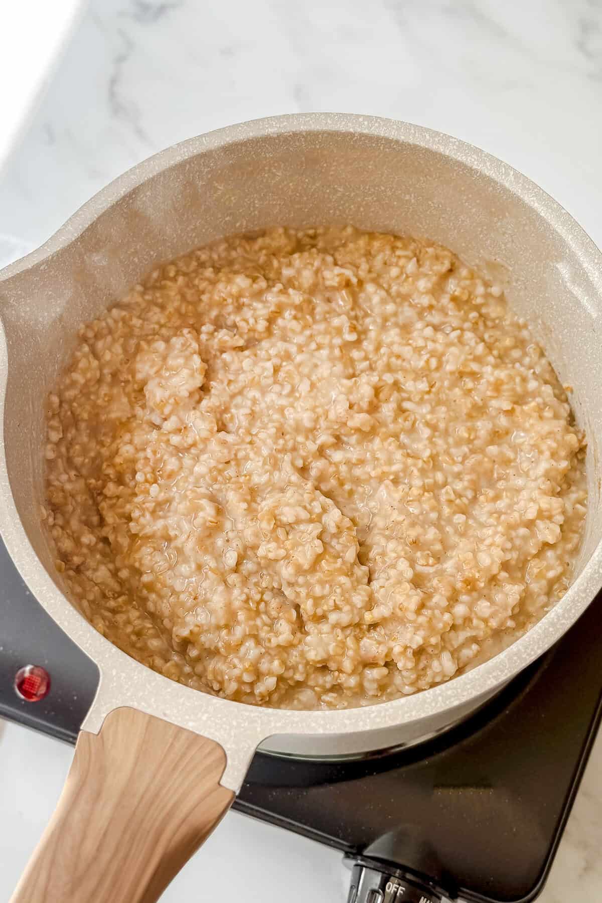 A beige saucepan filled with creamy Irish porridge sits on a black electric stovetop, its wooden handle visible against the light marble countertop in the background.