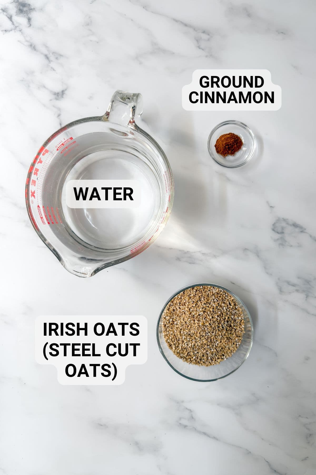 A glass measuring cup of water, a small glass bowl of ground cinnamon, and a bowl of Irish oats (steel cut oats) for Irish porridge sit on a white marble surface. Each item is clearly labeled with text.