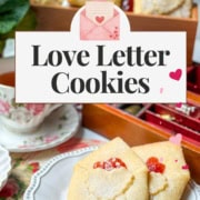 Two folded Love Letter Butter Cookies with a red jam center sit on a white plate, styled beside a teacup and a tray with more cookies. Text on image reads "Love Letter Cookies, 31Daily.com.