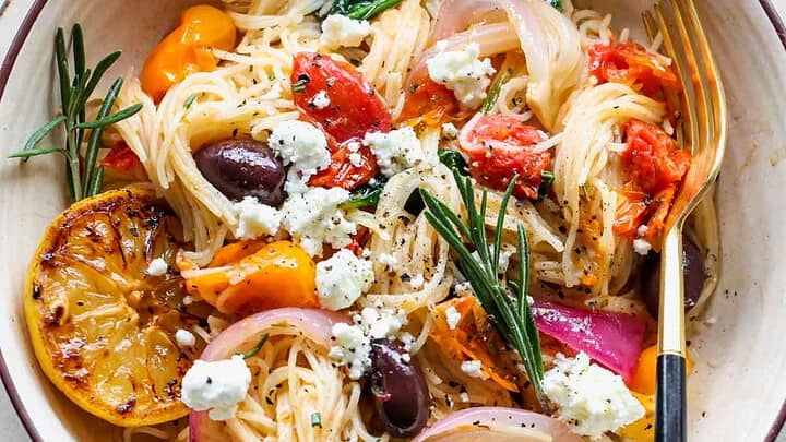 A bowl of pasta topped with cherry tomatoes, red onions, olives, spinach, grilled lemon, goat cheese, and herbs—perfect for February Mediterranean Diet Dinners—served with a fork.
