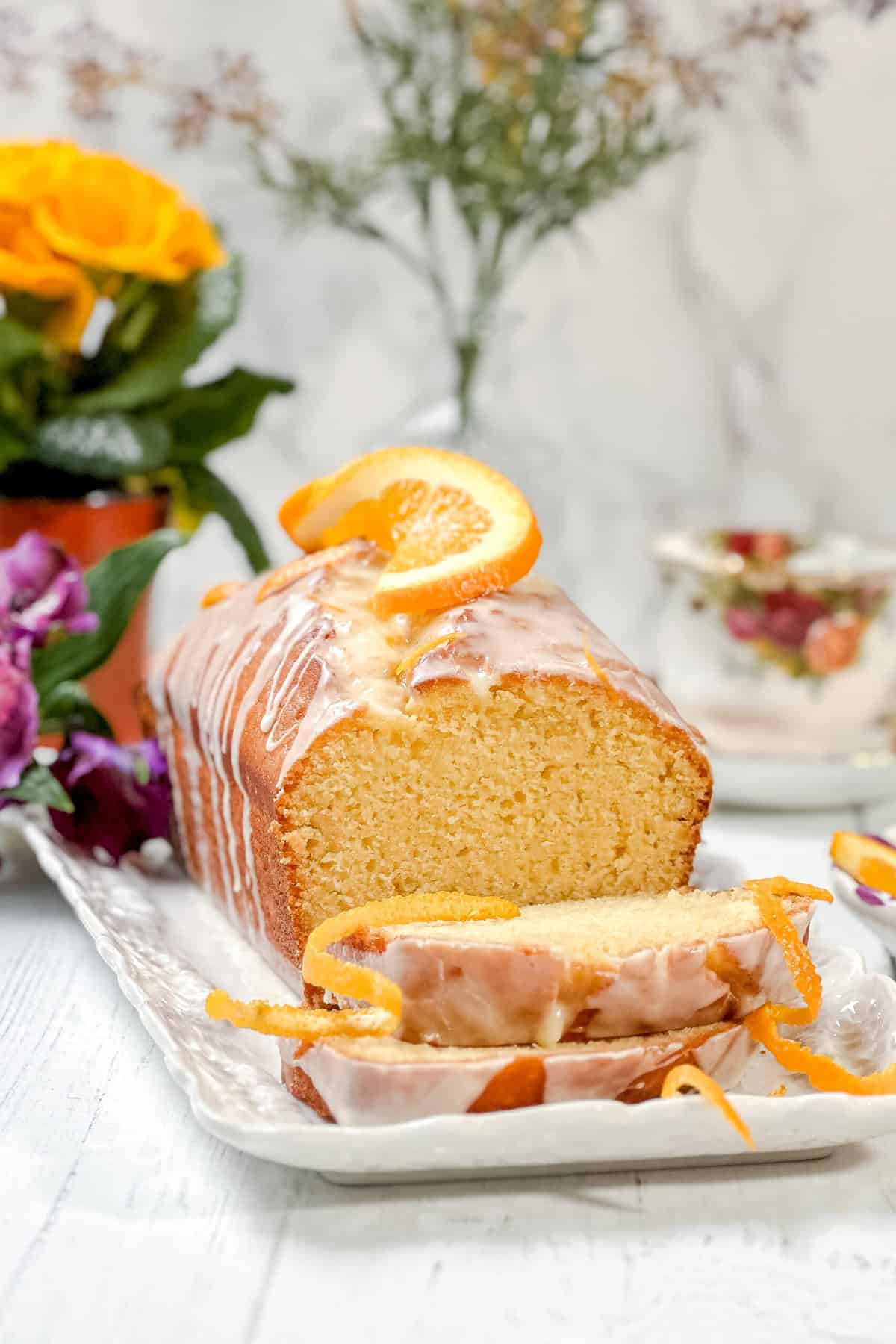 A loaf of glazed orange cake, partly sliced, is displayed on a rectangular white plate with an orange slice and zest as garnish. Flowers and a teacup are in the background, complementing the fresh appeal of this orange cake.