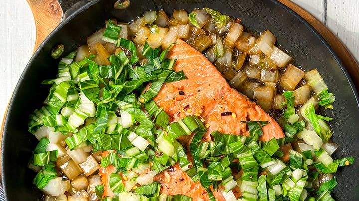 Top view of a salmon filet in a black nonstick skillet with onions and bok choy.