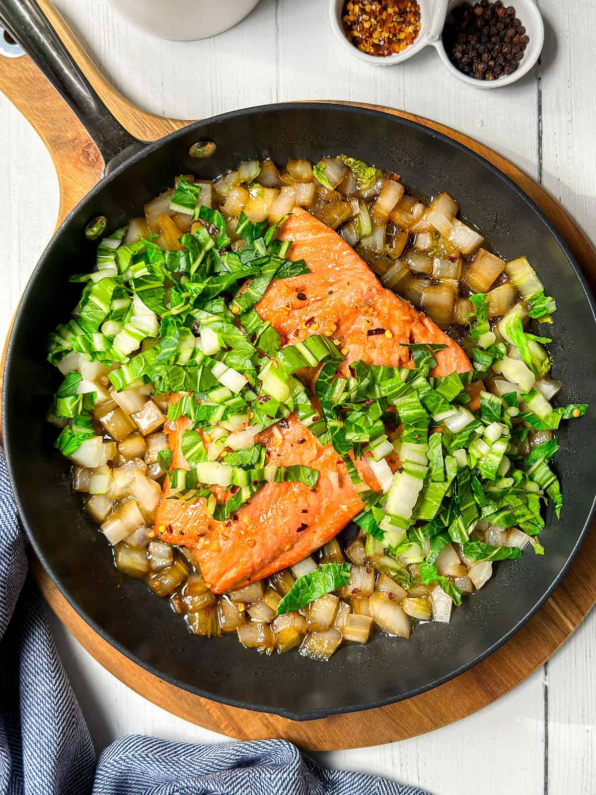 A skillet with two pan seared salmon fillets, chopped onions, and leafy greens cooking together, placed on a wooden board with spice bowls nearby.