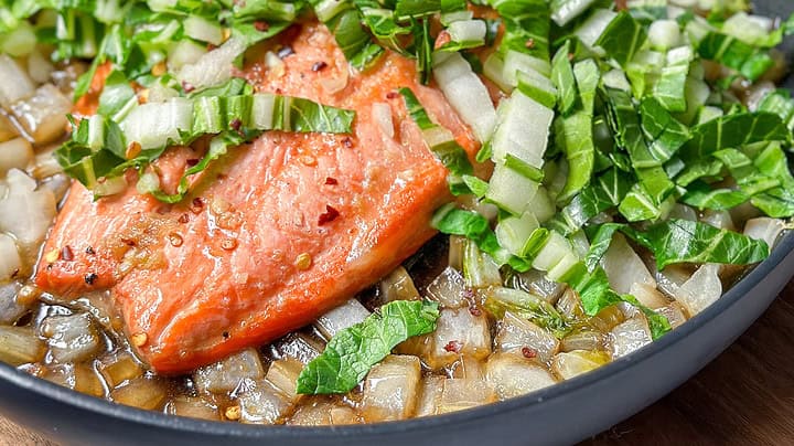 Side view of pan seared salmon with swiss chard simmered in a honey soy sauce in a black skillet.