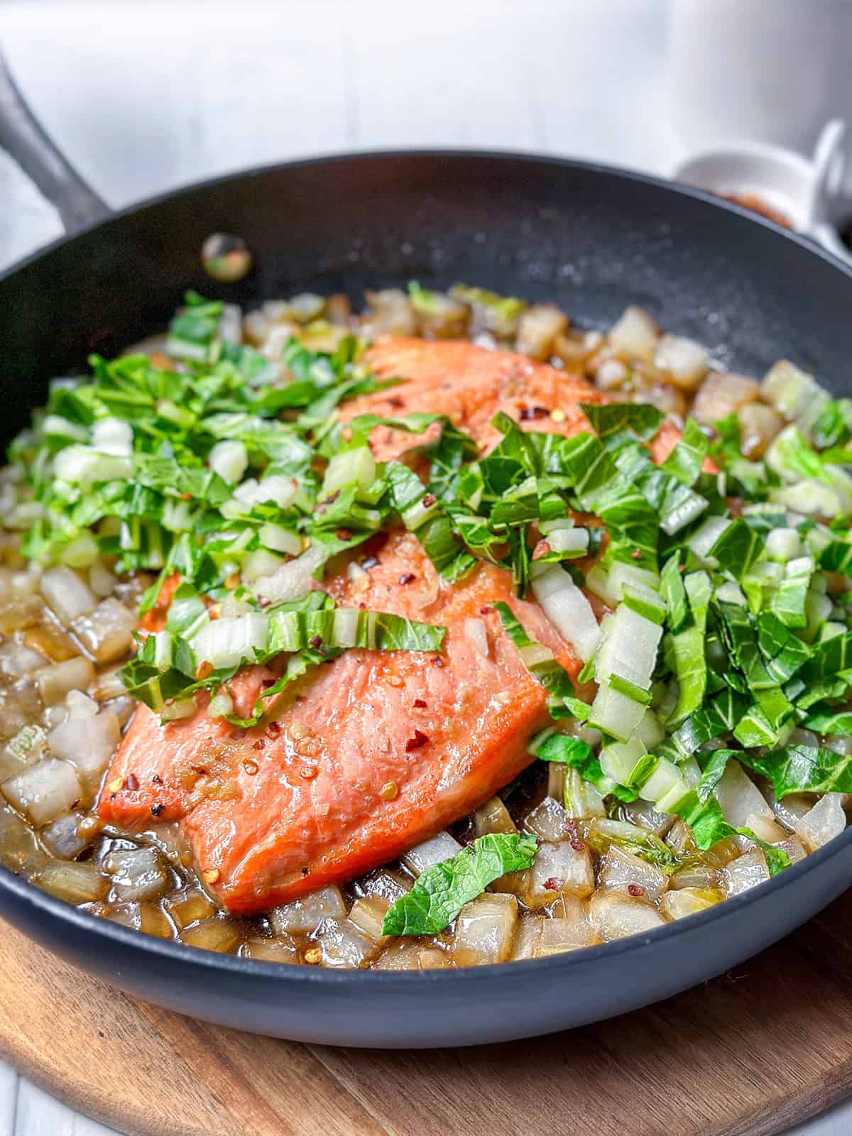Pan seared salmon fillet cooking in a skillet with chopped onions and greens on a wooden surface.