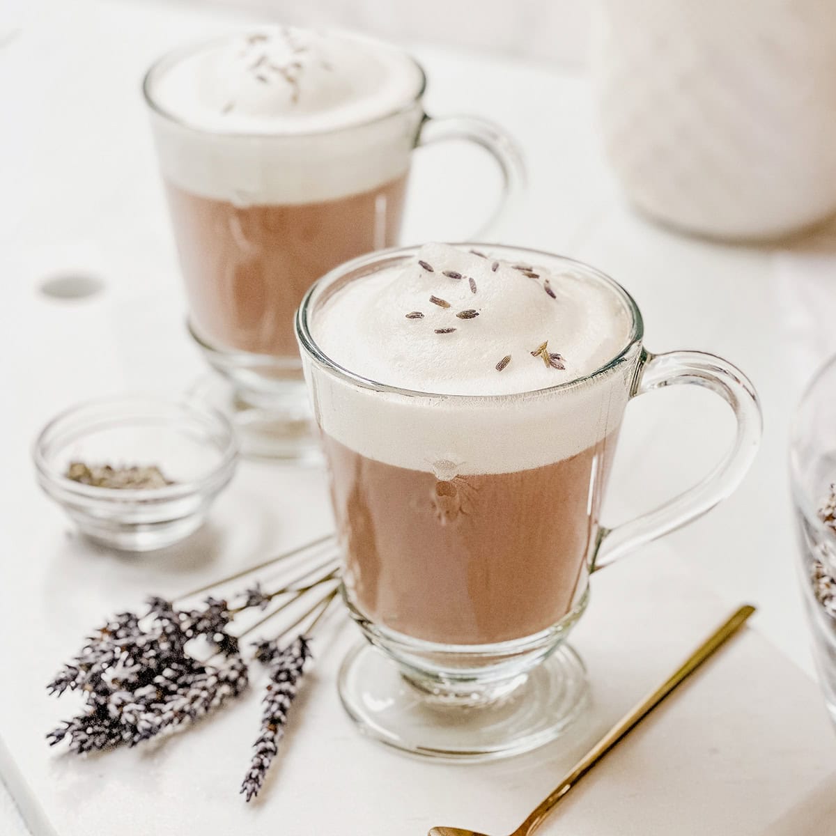 Two glass mugs filled with frothy lavender lattes, reminiscent of a lavender London fog, are topped with dried lavender buds and sit on a white surface next to a gold spoon and a small bowl of lavender flowers.