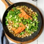 Pan seared salmon fillets, chopped onions, and greens are sizzling in a black skillet on a wooden board, with a blue-striped cloth nearby.