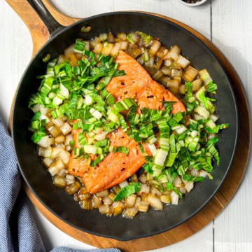Pan seared salmon fillets, chopped onions, and greens are sizzling in a black skillet on a wooden board, with a blue-striped cloth nearby.