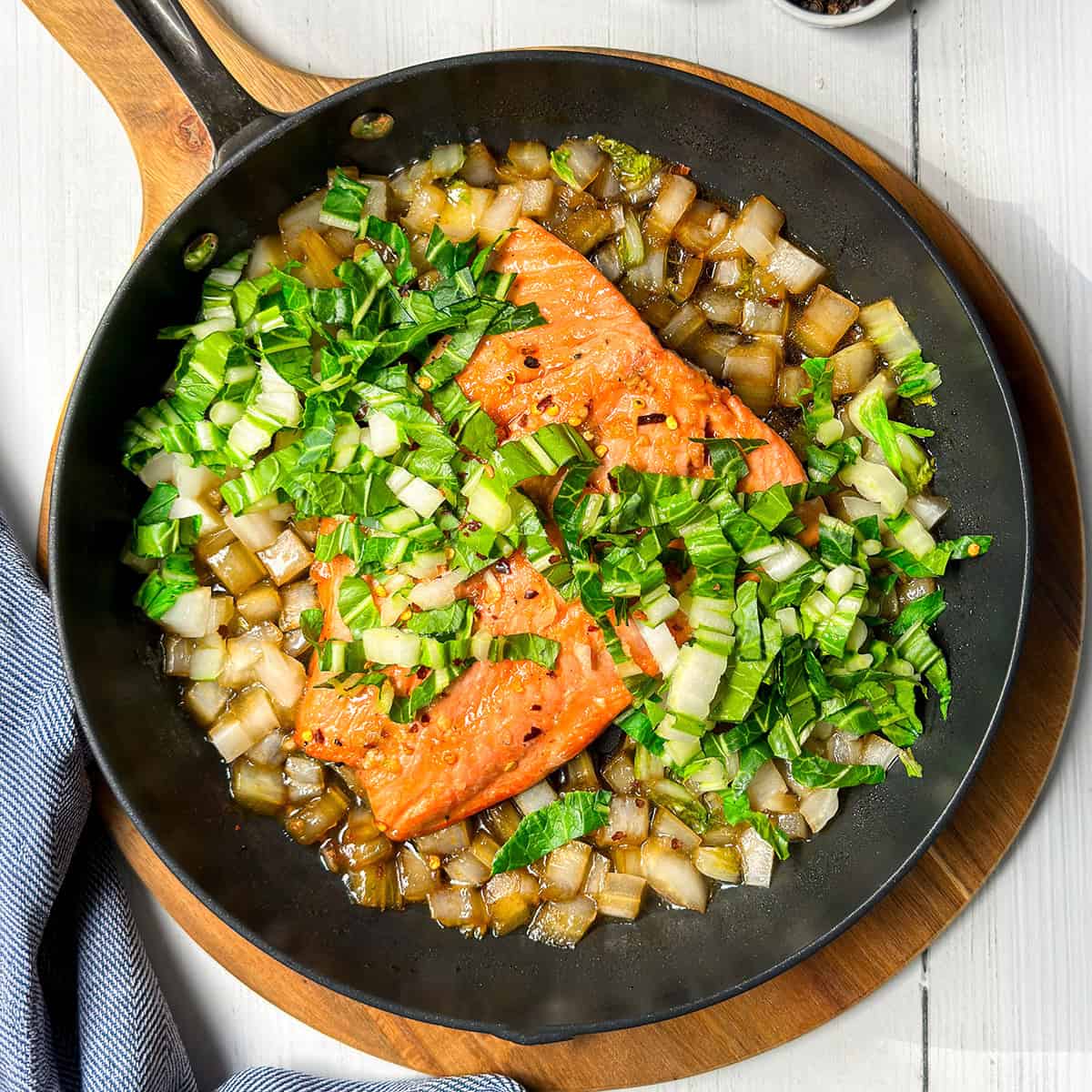 Pan seared salmon fillets, chopped onions, and greens are sizzling in a black skillet on a wooden board, with a blue-striped cloth nearby.