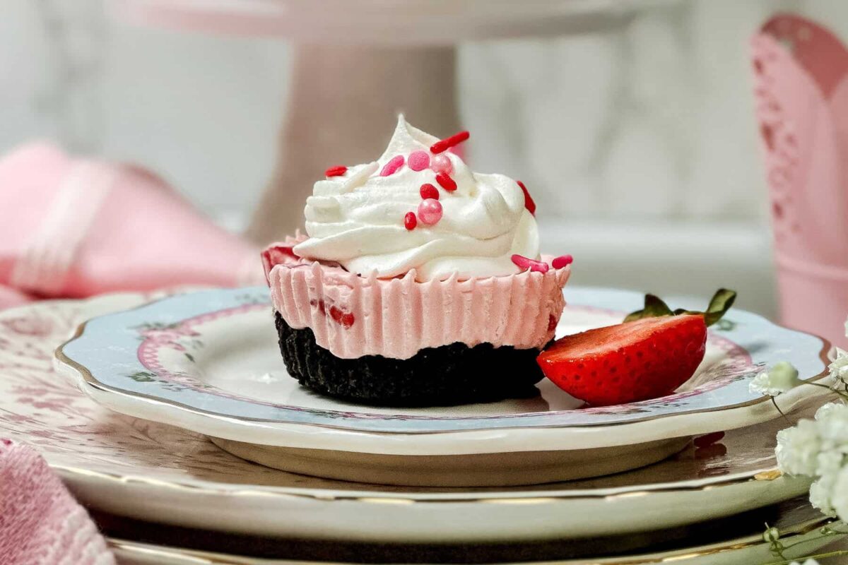 Horizontal side view of Strawberry No Bake Mini Cheesecake on a blue, pink, and white plate with whipped cream, sliced strawberries, and sprinkles.