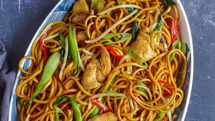 Oval plate of stir-fried Chinese New Year noodles with chicken, bell peppers, bean sprouts, and green vegetables, served with chopsticks on a dark surface.