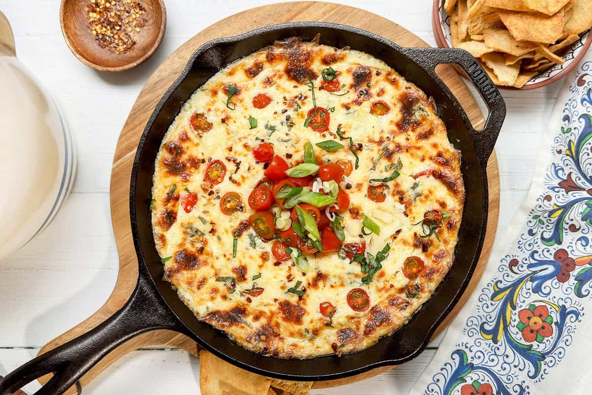 A skillet of caprese dip topped with melted cheese, cherry tomatoes, green onions, and herbs, placed on a wooden board with chips and a bowl of chili flakes nearby.