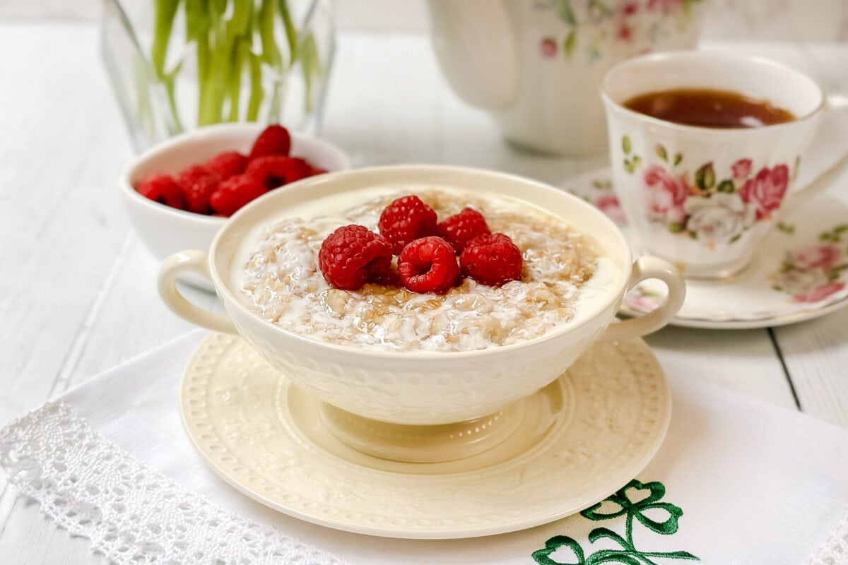 A bowl of Irish porridge topped with fresh raspberries sits on a white embroidered napkin, beside a bowl of raspberries and a floral teacup filled with tea. A vase with green leaves is in the background.