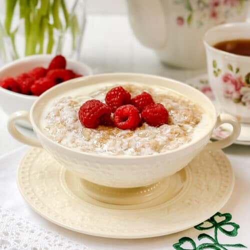 A bowl of Irish porridge topped with fresh raspberries sits on a white embroidered napkin, beside a bowl of raspberries and a floral teacup filled with tea. A vase with green leaves is in the background.