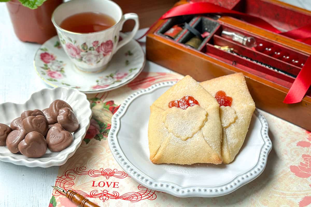 A plate with Love Letter Butter Cookies, a bowl of heart-shaped chocolates, a floral teacup with tea, and an open wooden box with a pen and red ribbon on a decorative surface.