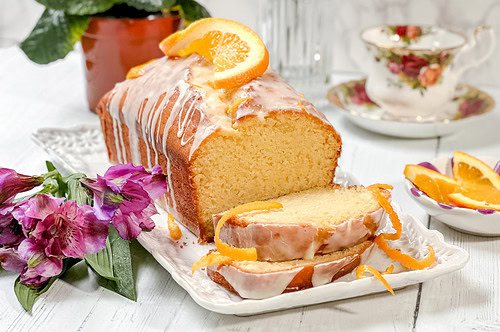 Side view of iced and sliced Orange Cake recipe with citrus zest, flowers, and orange slices served on a white rectangular serving dish.