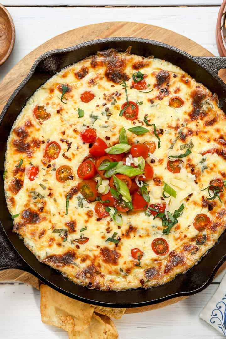 A warm caprese dip baked in a cast iron skillet, topped with cherry tomatoes, green onions, and herbs, served on a wooden board with chips and seasonings nearby.