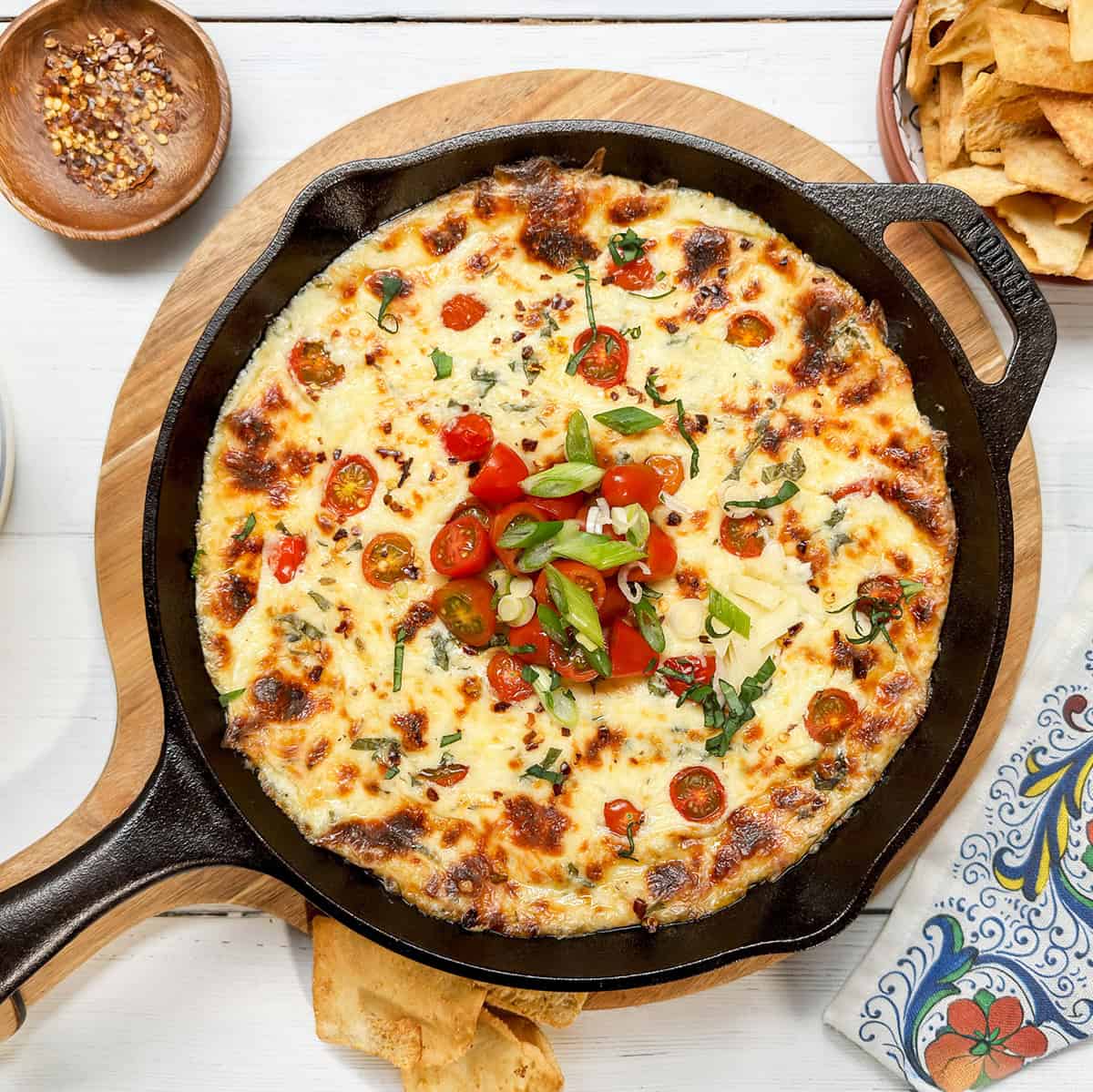 A warm caprese dip baked in a cast iron skillet, topped with cherry tomatoes, green onions, and herbs, served on a wooden board with chips and seasonings nearby.