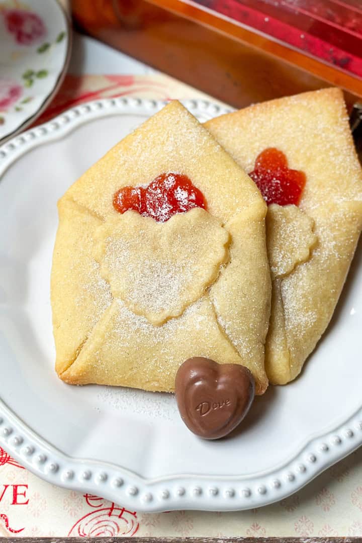 Two Love Letter Butter Cookies, envelope-shaped with heart-shaped jams and powdered sugar, sit on a white plate, accompanied by a heart-shaped chocolate.