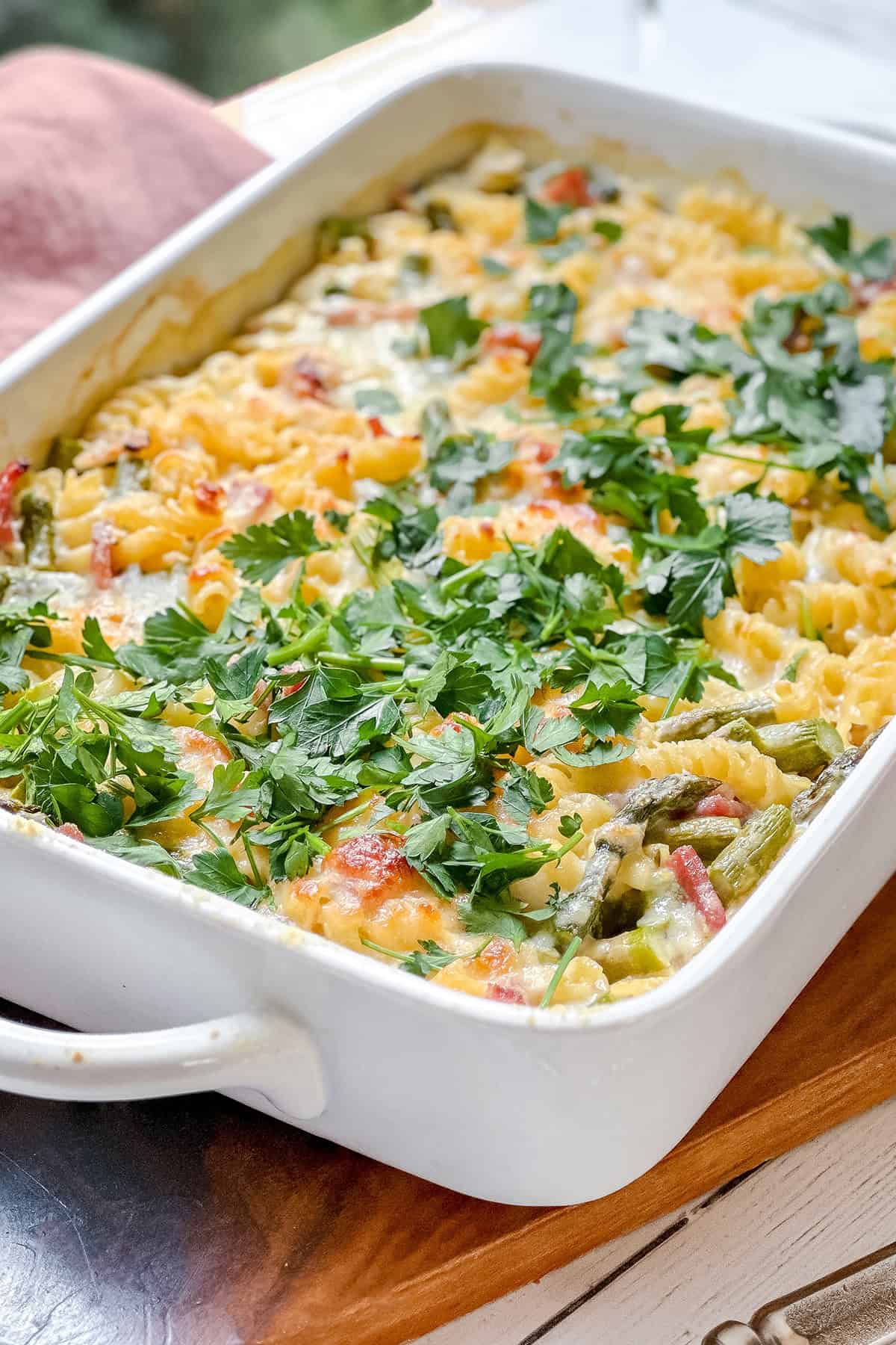 A baked pasta casserole topped with fresh parsley, featuring rotini pasta, vegetables, melted cheese, and savory ham&mdash;this Asparagus and Ham Casserole is served in a white rectangular baking dish.