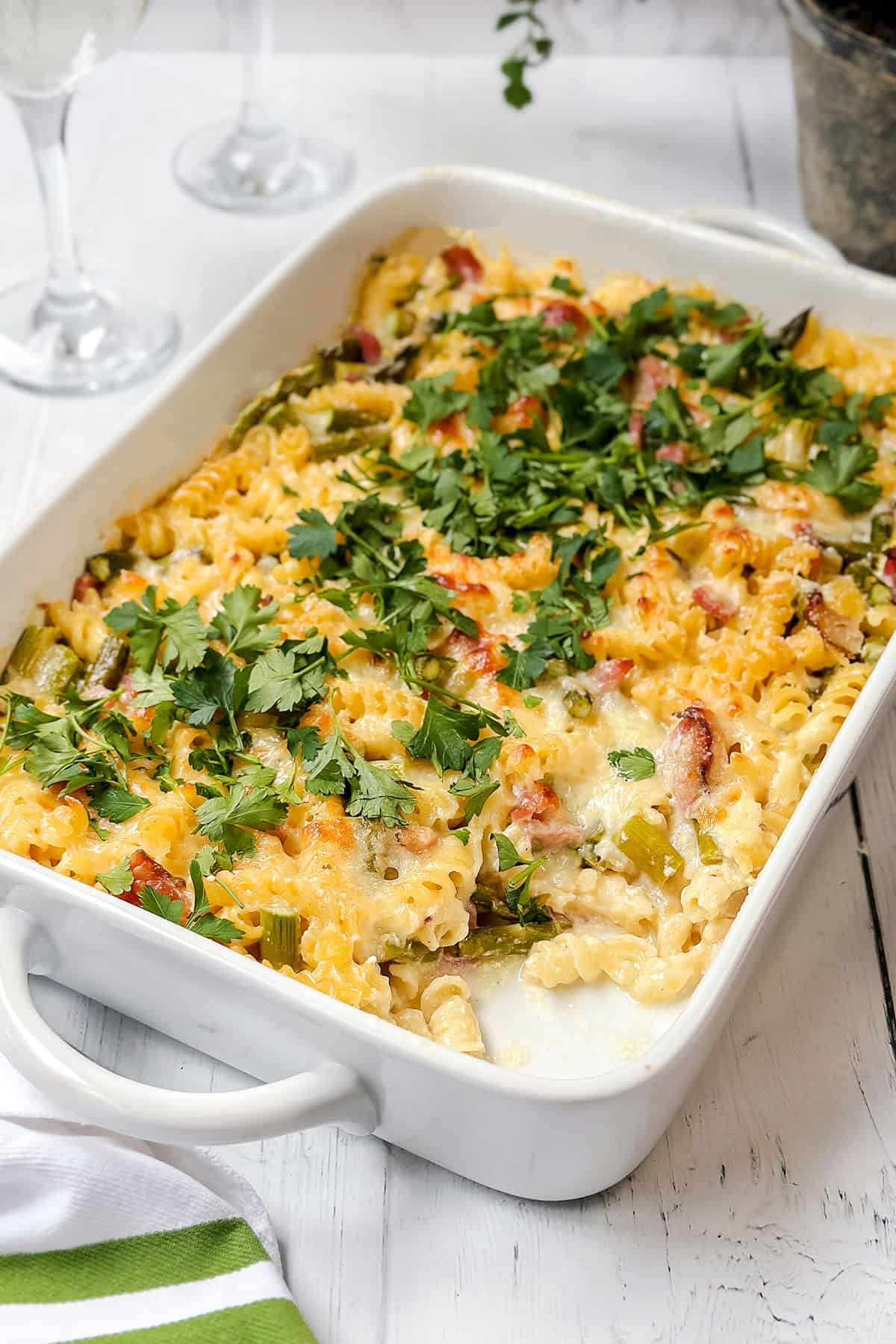A baked Asparagus and Ham Casserole with cheese, vegetables, and herbs, topped with fresh chopped parsley, in a white baking dish on a white wooden table.