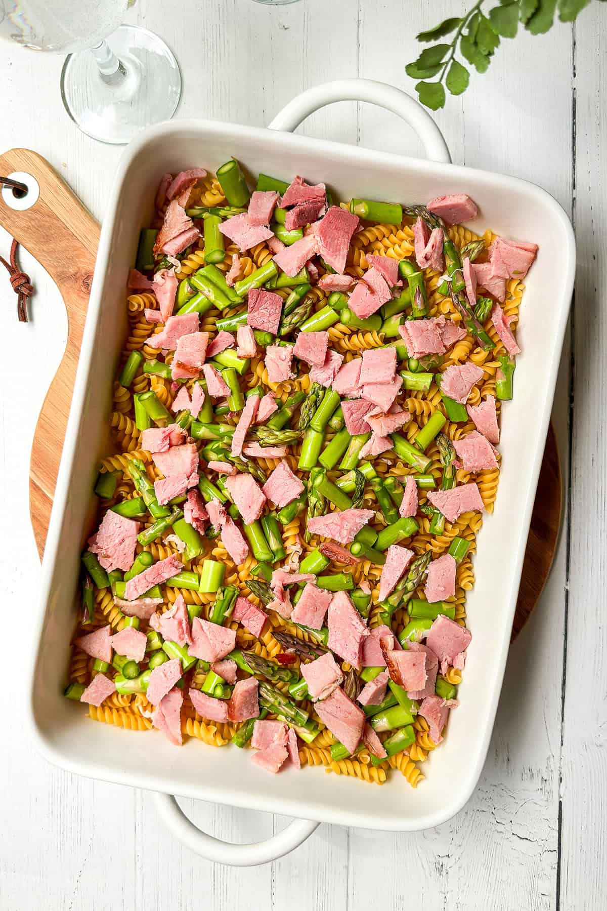 A white baking dish filled with uncooked rotini pasta, chopped asparagus, and pieces of ham on a white wooden table, ready to be baked into a delicious Asparagus and Ham Casserole.