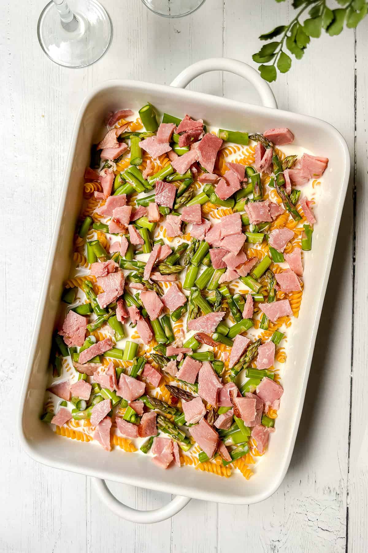 A rectangular white baking dish filled with uncooked pasta, chopped green asparagus, and pieces of pink ham-perfect for making a delicious Asparagus and Ham Casserole-rests on a white wooden surface.