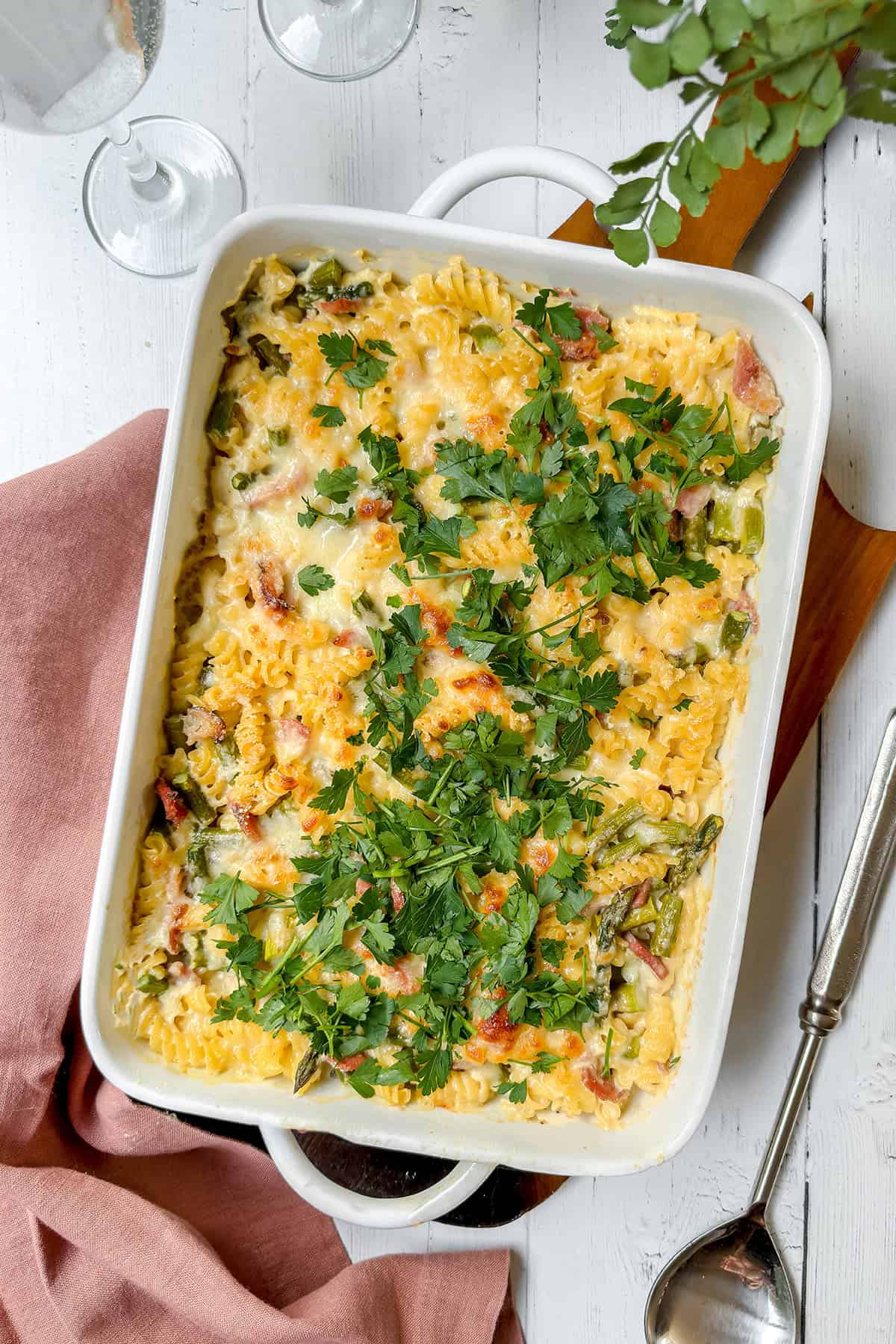 An Asparagus and Ham Casserole, baked with cheese and green herbs, is shown in a white dish on a wooden surface with a pink napkin and serving spoon nearby.