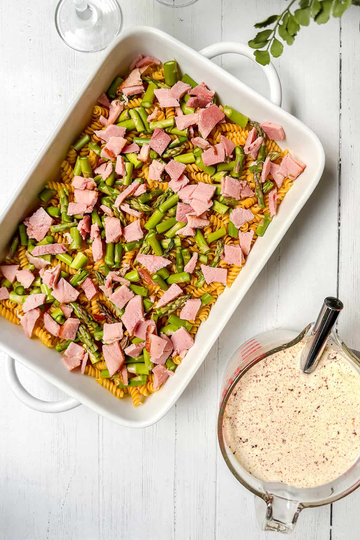 A white baking dish filled with dry rotini pasta, chopped ham, and asparagus sits on a white table next to a glass measuring cup containing homemade Alfredo sauce-perfect for making Asparagus and Ham Casserole.