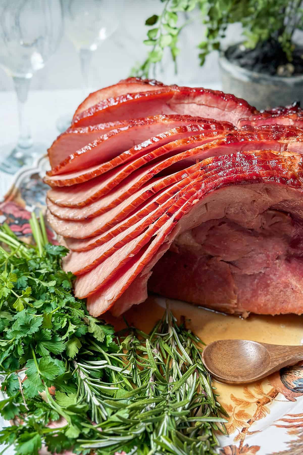 Sliced honey baked ham displayed on a table with fresh rosemary and cilantro, next to a wooden spoon and decorative greenery in the background.
